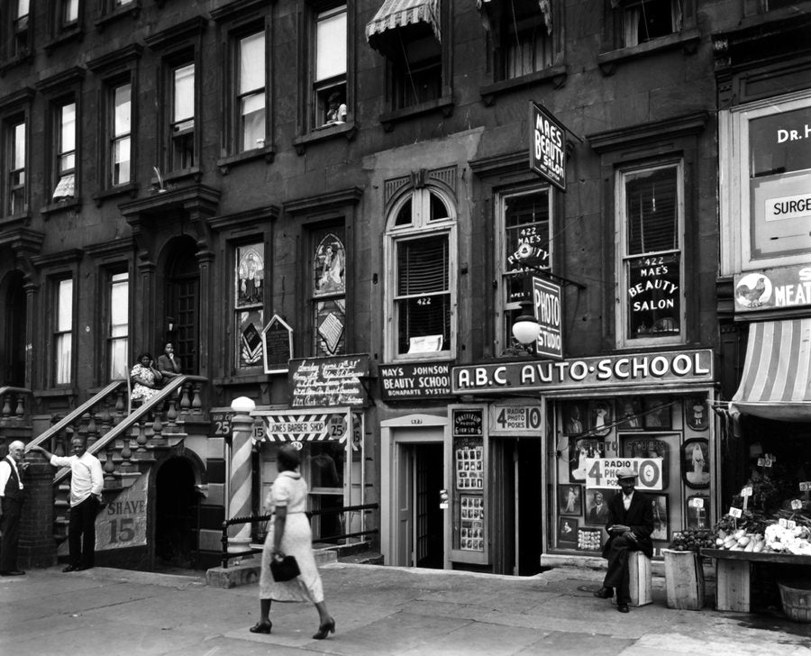 Lenox Avenue in Harlem in 1938: brownstone stoops, shop signs for a beauty salon and an auto school, a woman walking past.