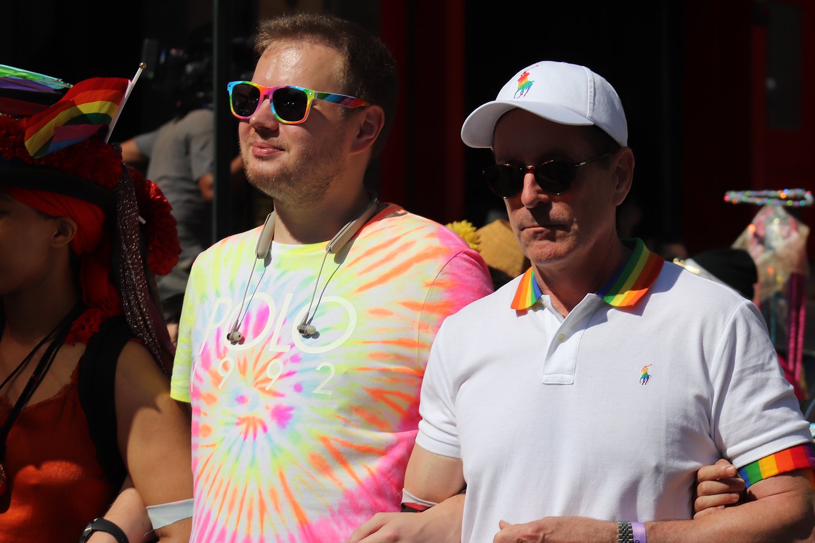 An older man in a white cap and a younger man in a tie-dye T-shirt march arm in arm in the sun.