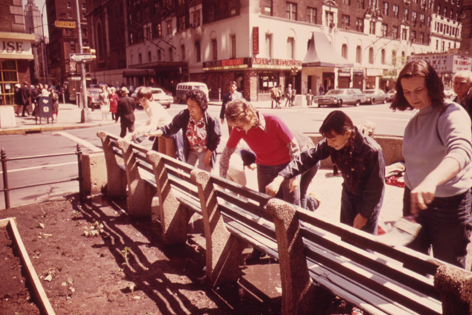 Neighbors of every age lean over a row of park benches with brushes, painting them on a sunny corner of Broadway.