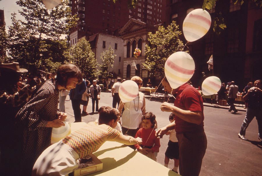 Neighbors of every age at a block party on a closed street, a boy blowing up balloons at a table.