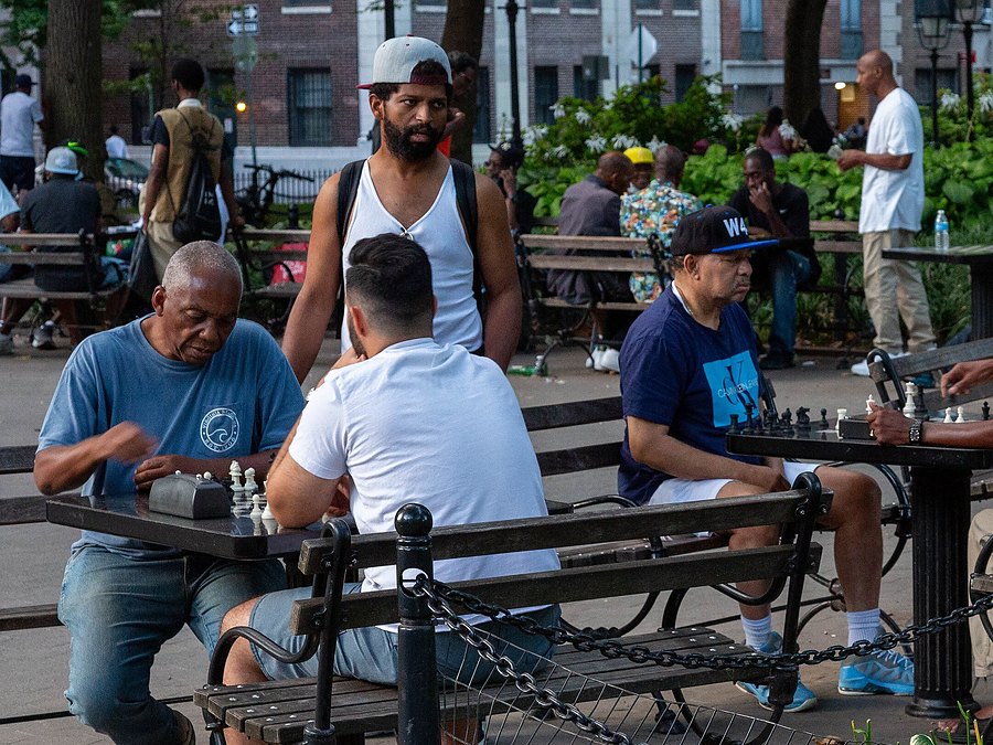 An older man plays chess against a younger man at a stone table in Washington Square Park while another young man looks on.