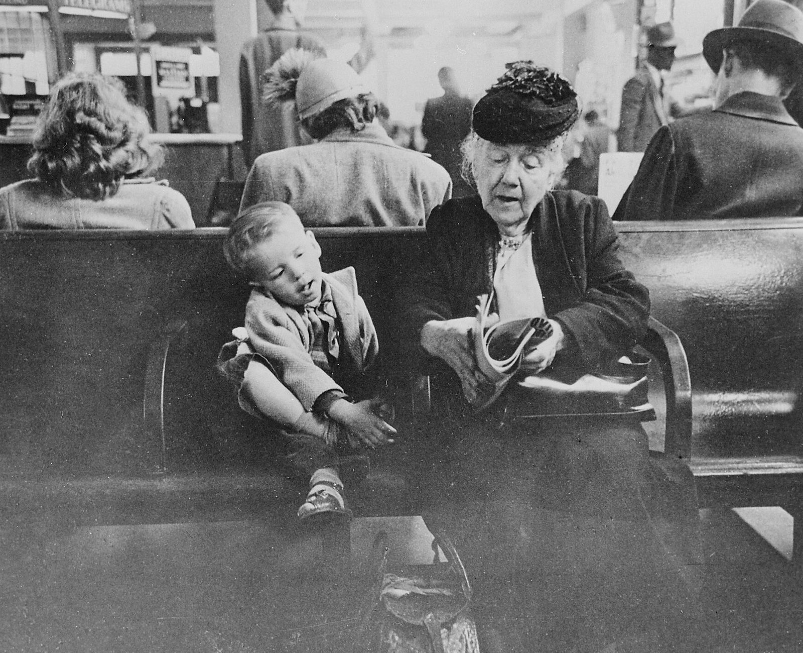 A grandmother in a feathered hat reads beside a small boy on a waiting-room bench, 1947.