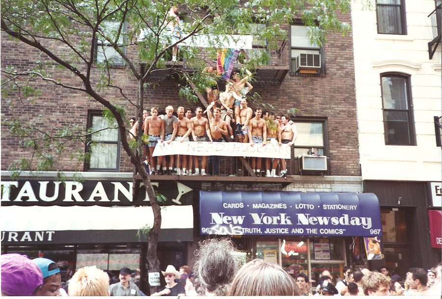Men on a fire escape hold a We Need Husbands banner over the crowd on Christopher Street, 1994.