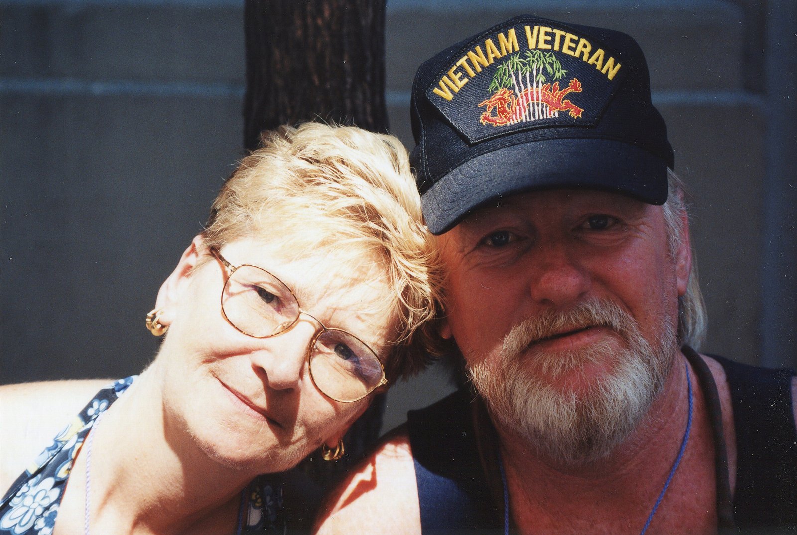 A woman in round glasses leans her head on a white-bearded man in a Vietnam Veteran cap, both smiling, at Pride in Greenwich Village.