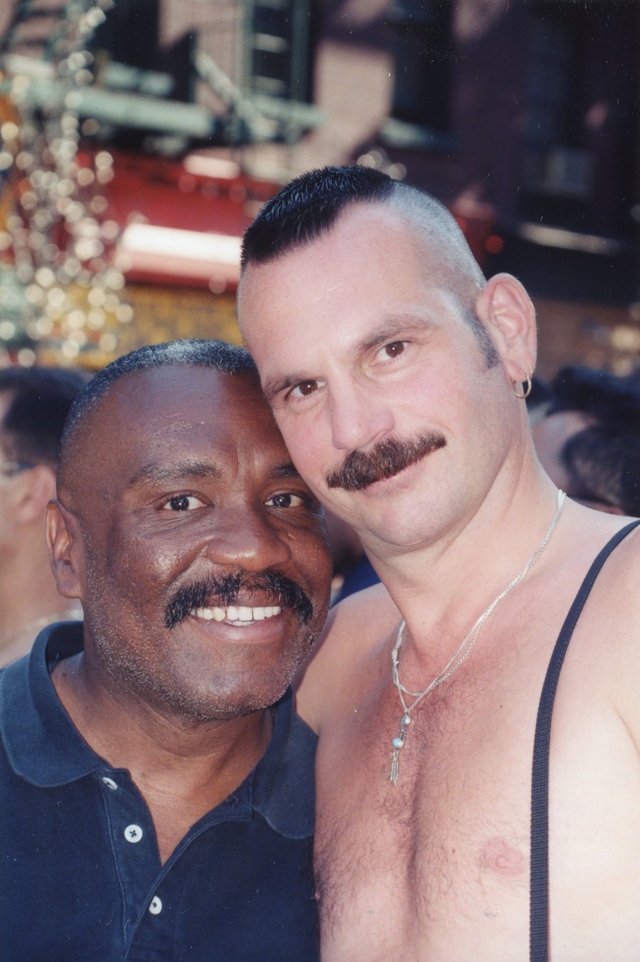 Two older men with moustaches stand close and smile at Pride, 1997.
