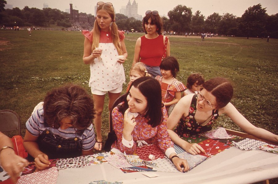Women and girls of several ages gather round a patchwork quilt on the grass in Central Park, stitching it together.