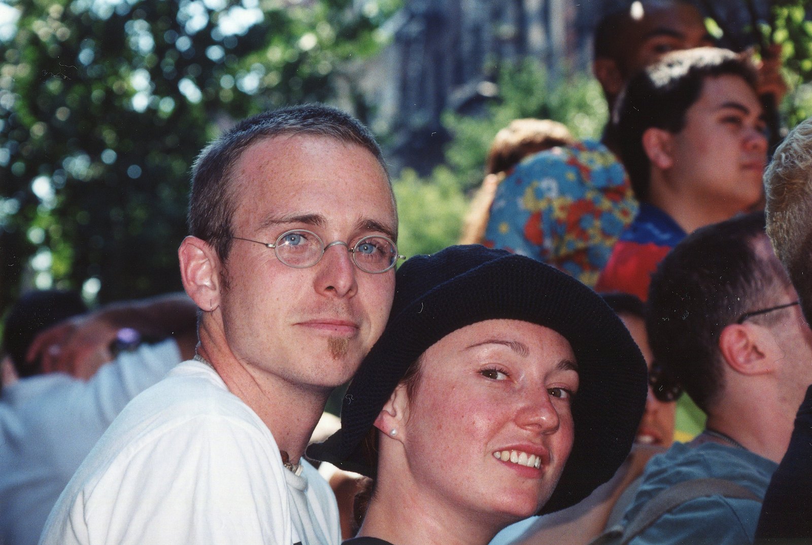 Two marchers, one in round glasses and one in a black hat, lean together and smile at the camera in the Pride crowd.