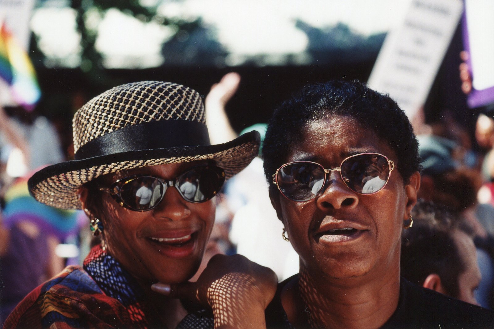 Two women in sunglasses, one in a woven straw hat, smile together in the crowd at Pride in Greenwich Village.