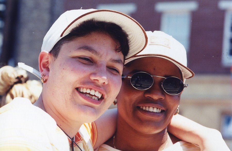 Two women in white caps hug cheek to cheek and laugh at the camera at Pride.